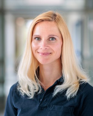 Blonde Frau mit langen Haaren und blauen Augen, trägt ein schwarzes Polo-Shirt mit dem Logo "srh". Hintergrund unscharf.