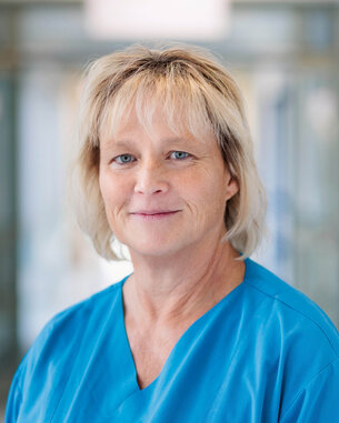 Eine Frau mit blonden Haaren trägt ein blaues medizinisches Outfit und lächelt leicht in die Kamera.