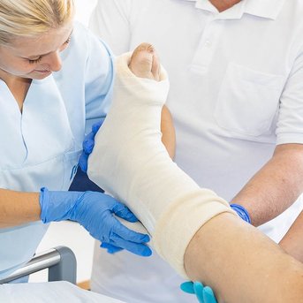 Zwei Pflegekräfte verbinden einen verletzten Fuß eines Patienten, während ein Arzt Anweisungen gibt. Der Patient liegt auf einem Bett in einem hellen Krankenhauszimmer.