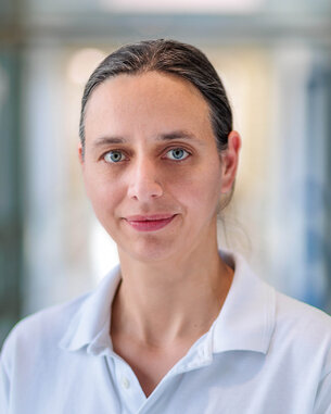 Eine Frau mit langen, glatten Haaren und blauen Augen trägt ein weißes Oberteil. Der Hintergrund ist unscharf und hell.