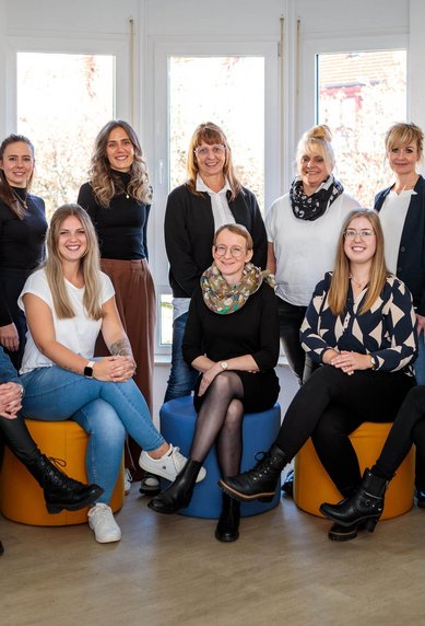 Eine Gruppe von zwölf Frauen sitzt auf farbigen Hockern in einem hellen Raum. Sie tragen verschiedene formelle Outfits und lächeln in die Kamera.