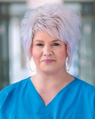 Frau mit blonden, lockigen Haaren trägt ein blaues Oberteil. Sie steht in einem hellen, modernen Raum und schaut direkt in die Kamera.