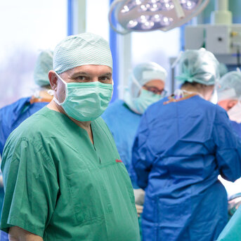 Ein Chirurg in grüner OP-Kleidung und Maske steht im Vordergrund, während ein Team von Fachleuten hinter ihm an einem chirurgischen Eingriff arbeitet.