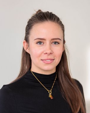 Eine Frau mit langen, braunen Haaren trägt ein schwarzes Oberteil und eine goldene Halskette. Sie schaut direkt in die Kamera.