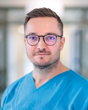 Ein Mann in einem blauen medizinischen Kittel mit Brille steht in einem hellen, unscharfen Hintergrund. Er hat einen neutralen Gesichtsausdruck und trägt ein Logo auf der linken Brustseite.
