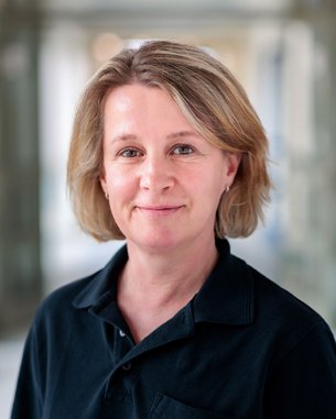 Frau mit blonden, schulterlangen Haaren trägt ein schwarzes Polohemd. Sie steht vor einem unscharfen Hintergrund.