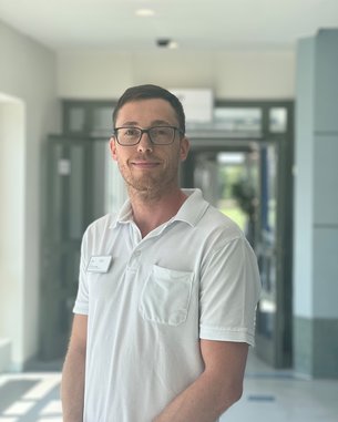 Ein Mann mit Brille trägt ein weißes Polohemd und steht in einem hellen Flur. Im Hintergrund sind Türen und Fenster sichtbar.