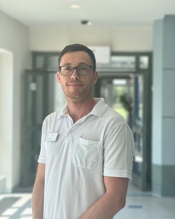 Ein Mann mit Brille trägt ein weißes Polohemd und steht in einem hellen Flur. Im Hintergrund sind Türen und Fenster sichtbar.