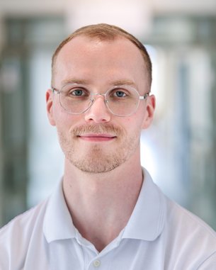 Ein junger Mann mit Brille und kurzem Haar, der in einem hellen, modernen Raum steht und in die Kamera schaut.